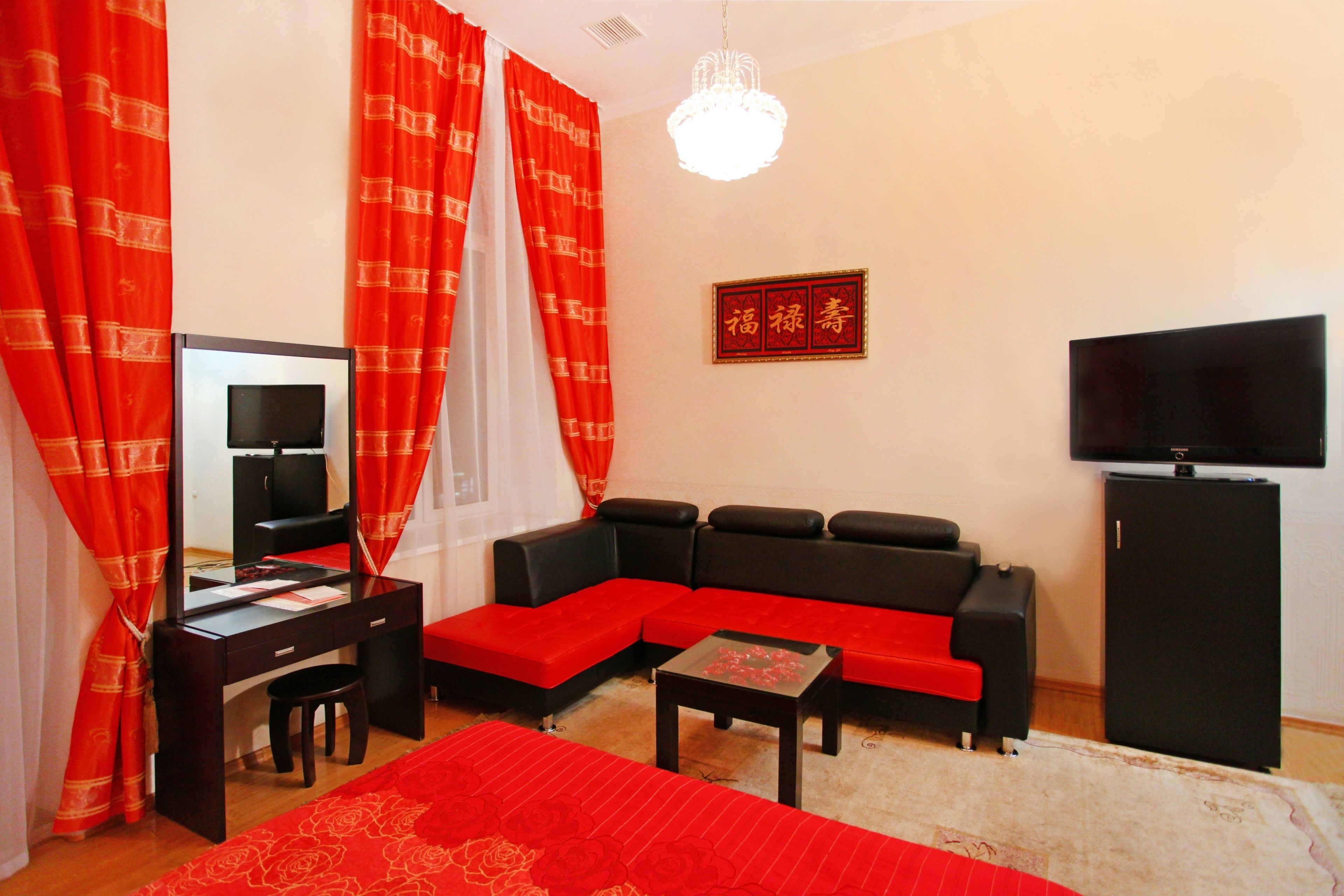 A room with a black sofa, a red and white patterned curtain, a small table, a television, and a refrigerator.