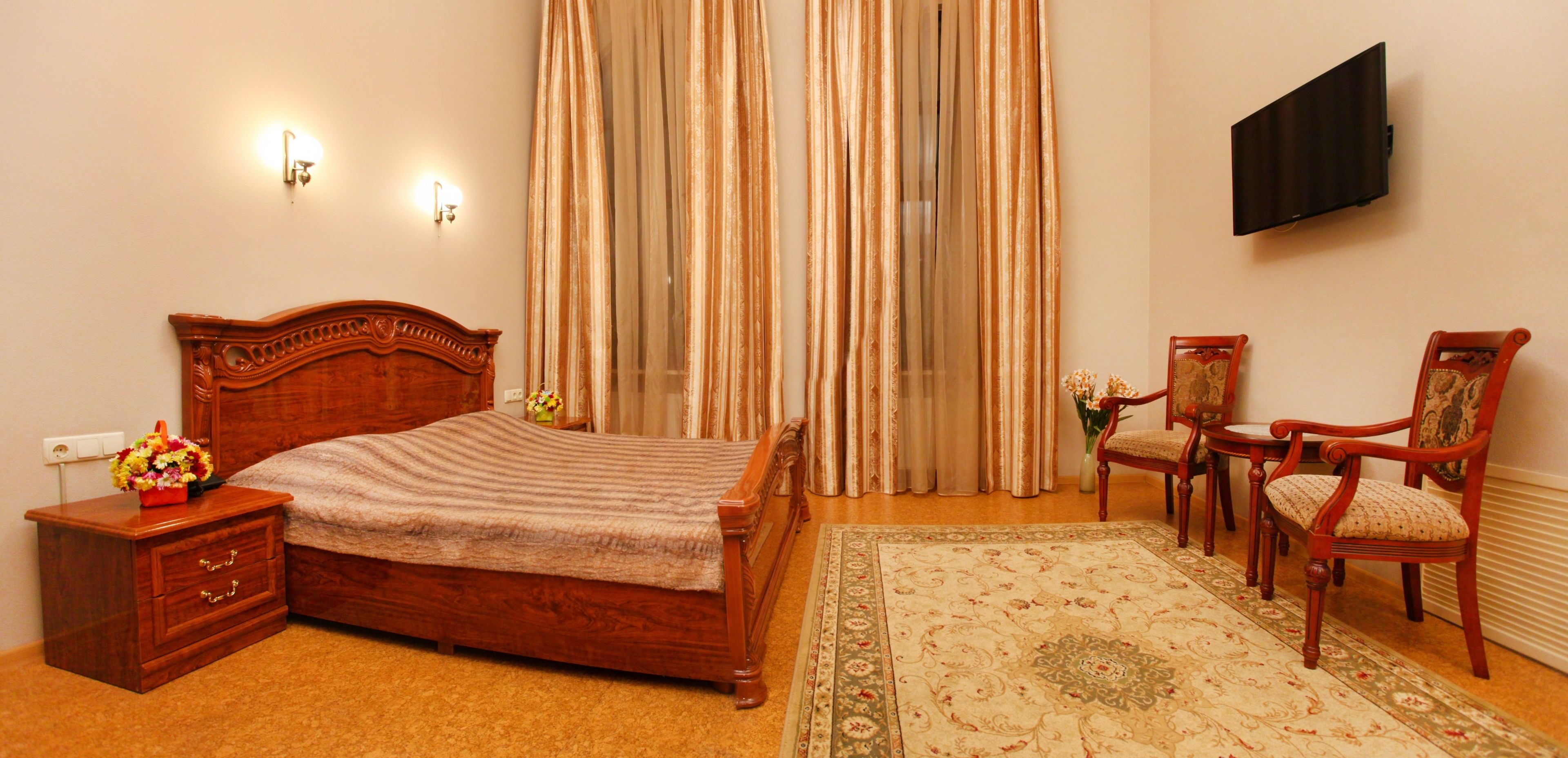 Un dormitorio con cama de madera, una mesita de noche con un jarrón de flores, una zona de estar con dos sillones, un televisor montado en la pared y un amplio ventanal con cortinas.