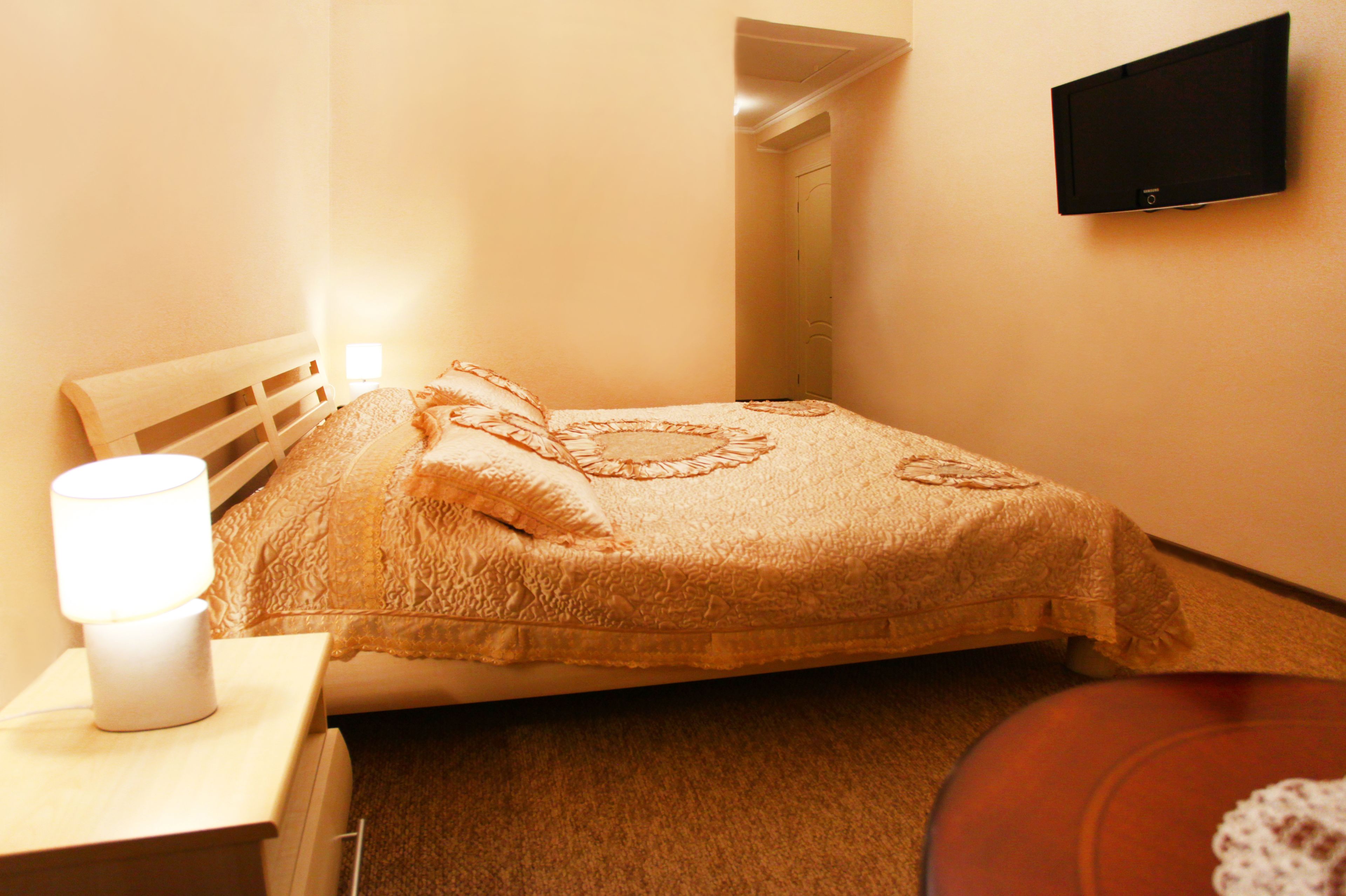 A bedroom with a bed, a bedside table with a lamp, a flat-screen TV mounted on the wall, and a door leading to another room.