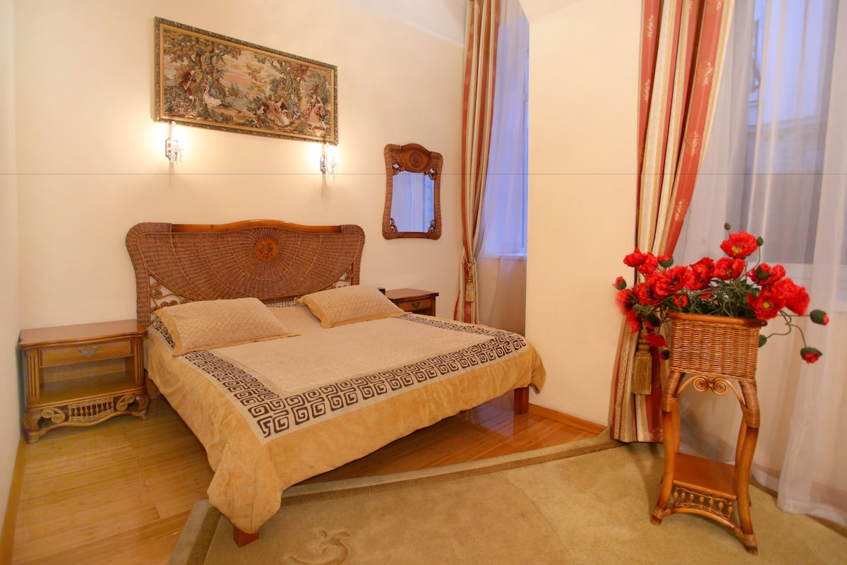 Un dormitorio con cama, mesita de noche, cómoda, espejo, ventana con cortinas y un jarrón con flores.