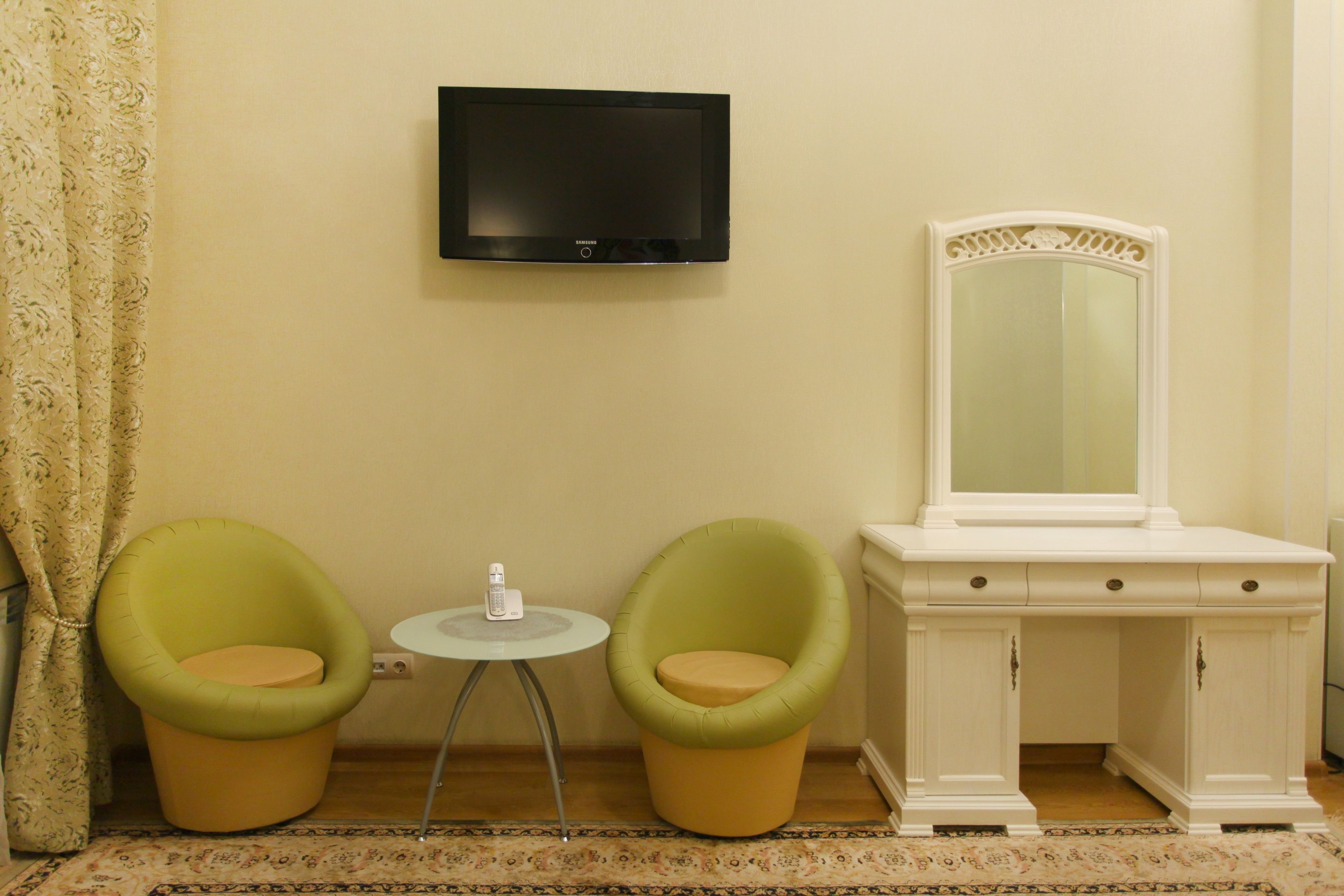 A room with two green chairs, a small round table, a white dresser with a mirror, and a flat-screen TV mounted on the wall.
