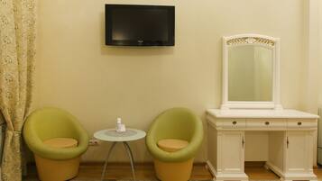 A room with two green chairs, a small round table, a white dresser with a mirror, and a flat-screen TV mounted on the wall.