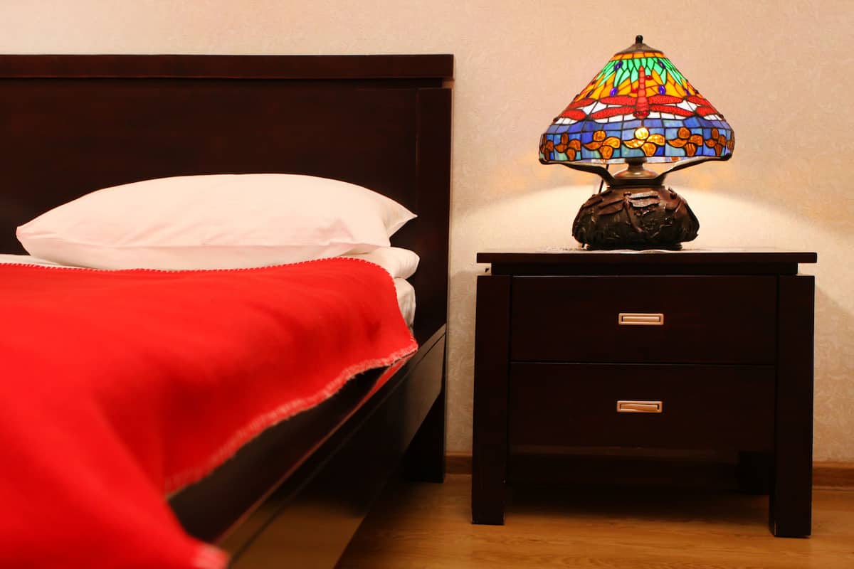 A bed with a white pillow and a red blanket, a small wooden nightstand with a stained glass lamp, and a wall with a subtle pattern.