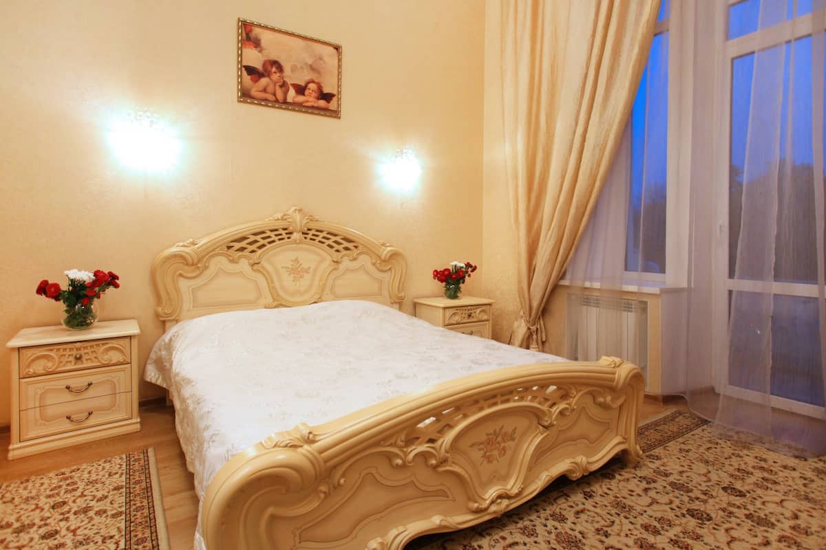 A bedroom with a large bed, two bedside tables, a nightstand, a window with curtains, and a wall-mounted picture.