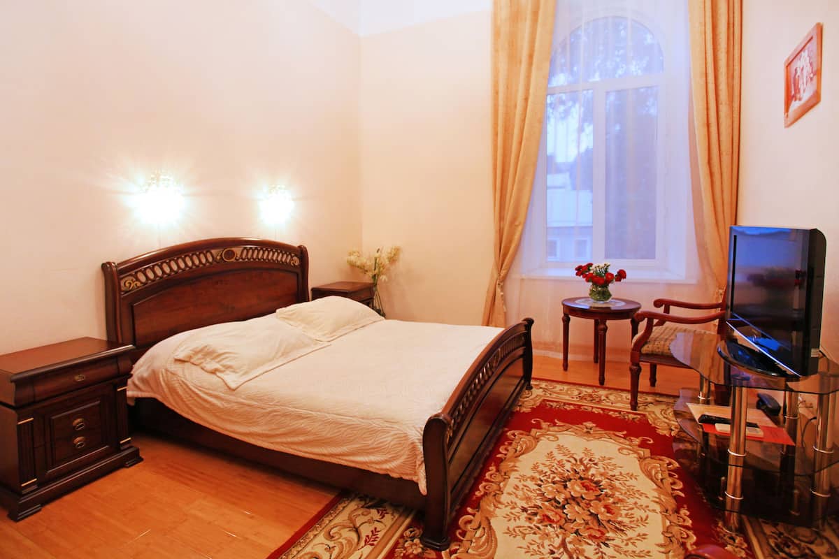 A bedroom with a wooden bed, bedside tables, a TV on a glass stand, a small table with a vase, and a window with curtains.