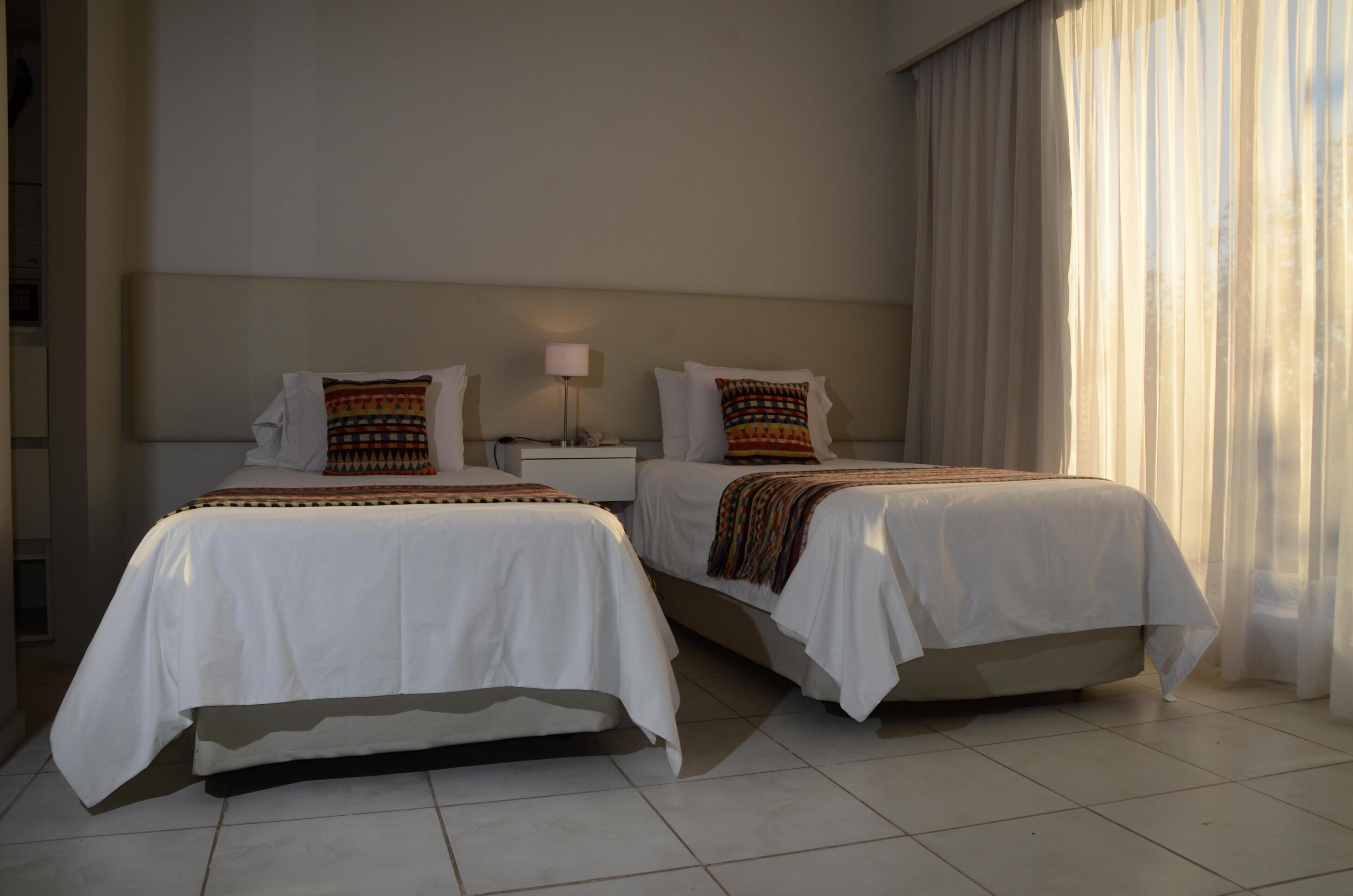 A hotel room with two beds, a nightstand, a lamp, and a window with curtains.