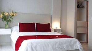A neatly made bed with white and red bedding, a bedside table with a lamp, and a vase with flowers. - Apart Via 51 (La Plata)