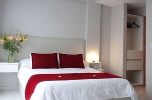 Una cama bien hecha con ropa de cama blanca y roja, una mesita de noche con una lámpara y un jarrón con flores.