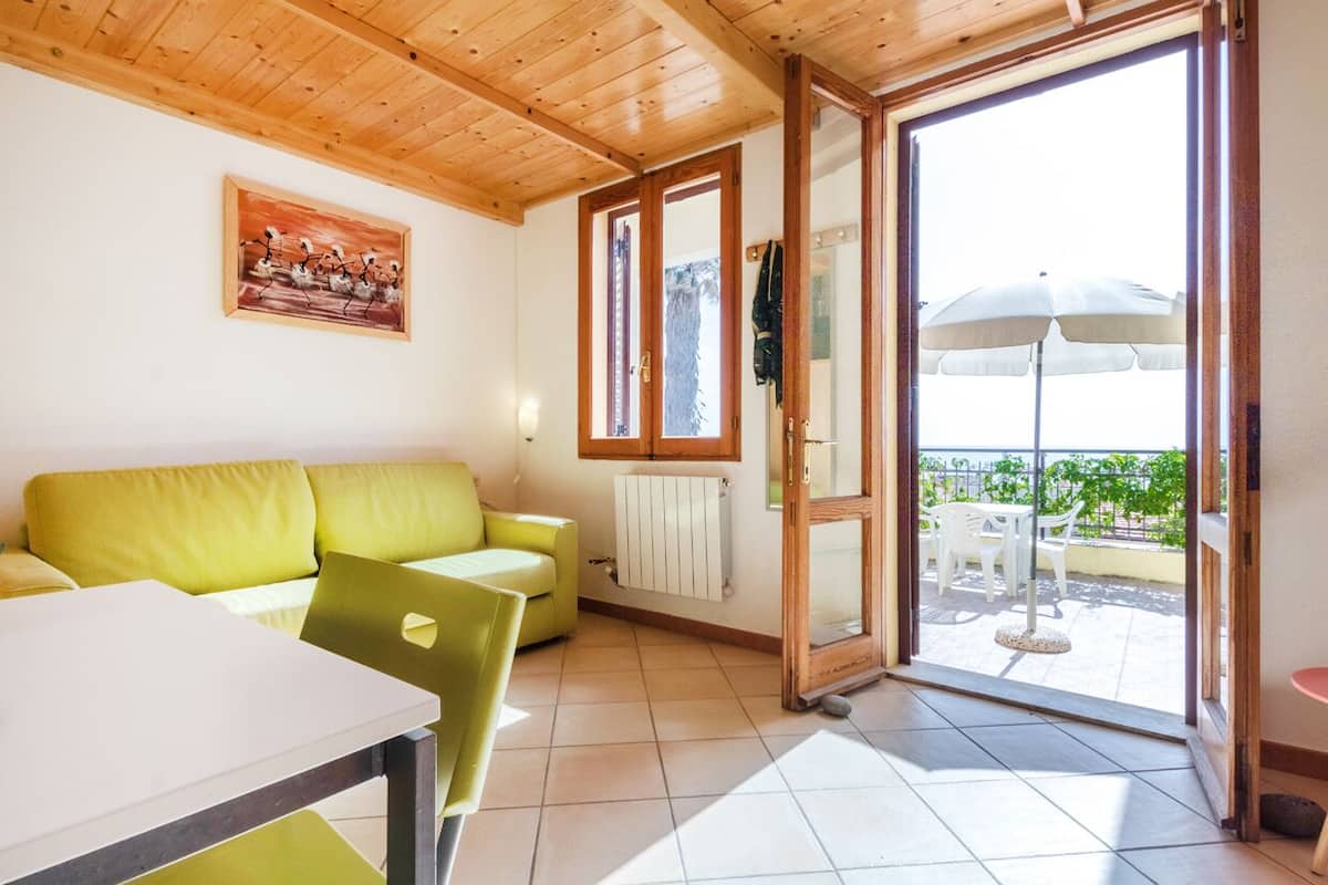 A room with a wooden ceiling, a green sofa, a white table, and a view through a glass door to an outdoor patio with umbrellas.