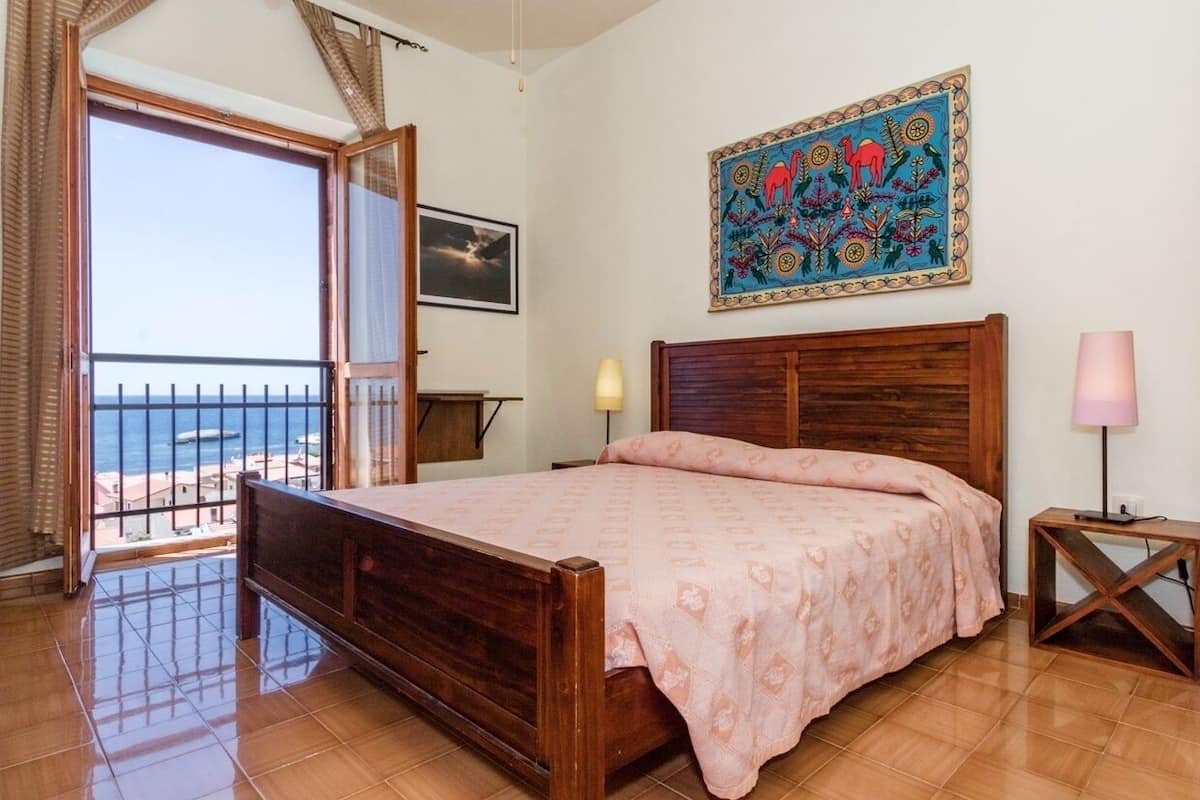 A bedroom with a wooden bed, a balcony with a view, a painting on the wall, and a lamp on a bedside table.