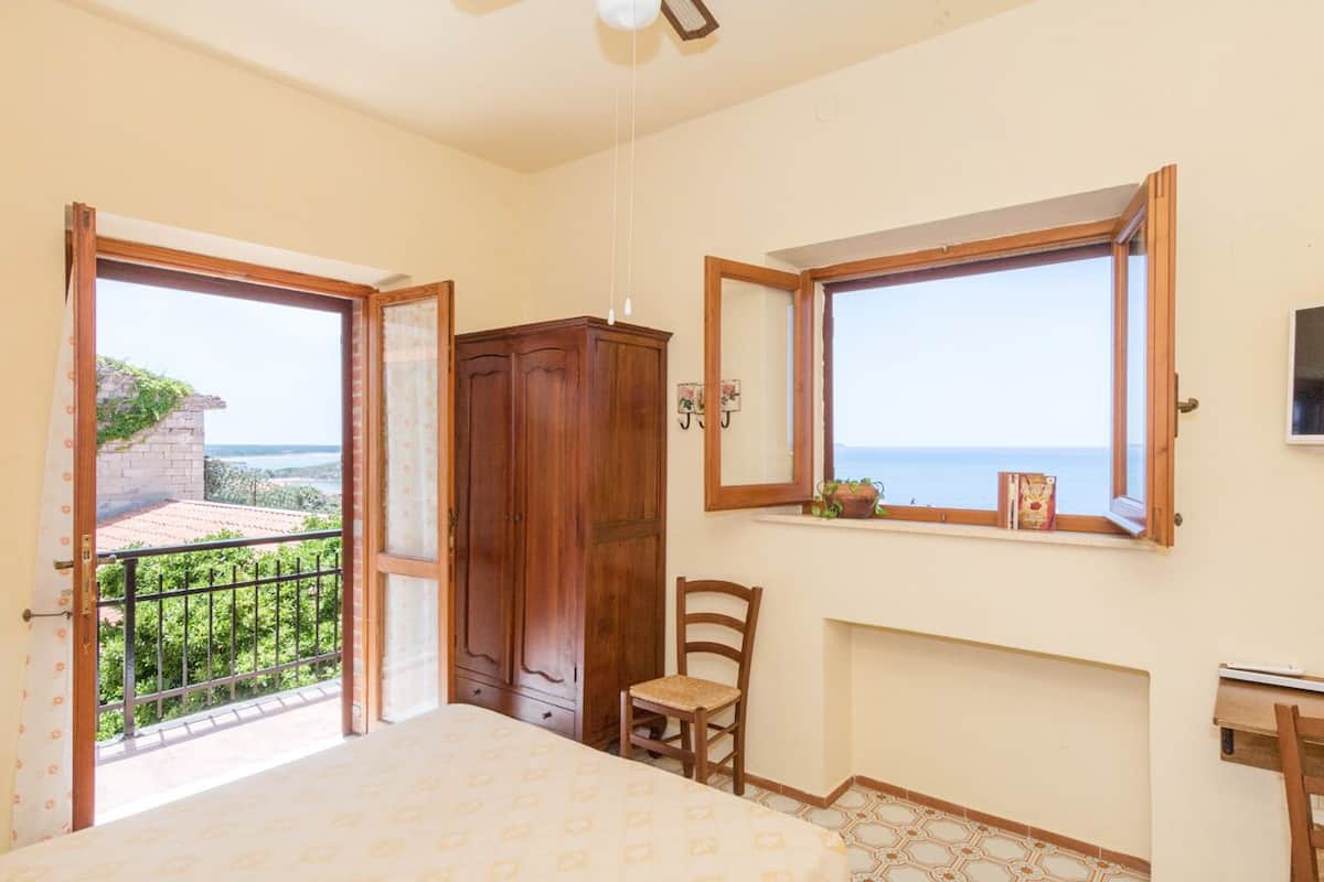 A bedroom with a bed, a wooden wardrobe, a chair, a window with a view of the sea, and a small table.