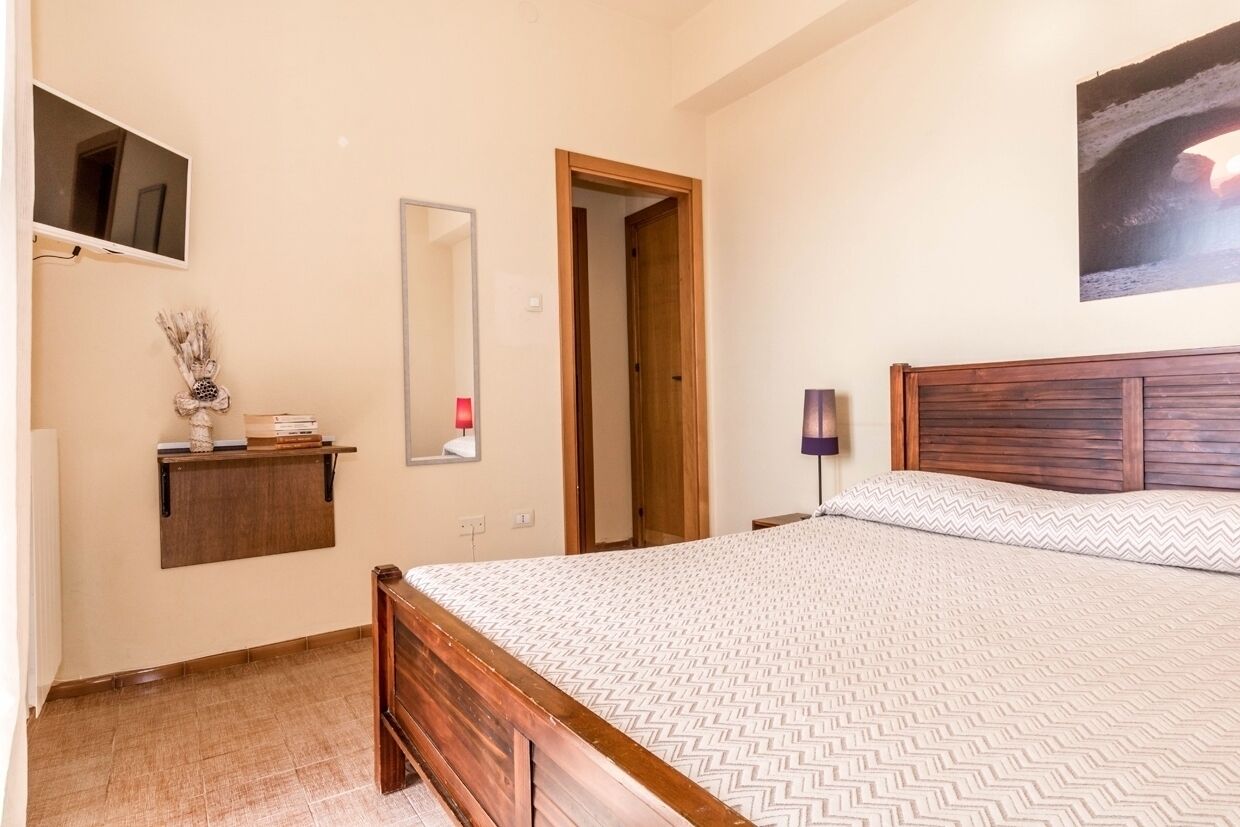A bedroom with a wooden bed, a bedside table, a mirror, a TV mounted on the wall, and a door leading to another room.