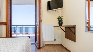 A bedroom with a bed, a TV mounted on the wall, a potted plant, and a view of the sea through an open door. - Mare Lughente (Cuglieri)