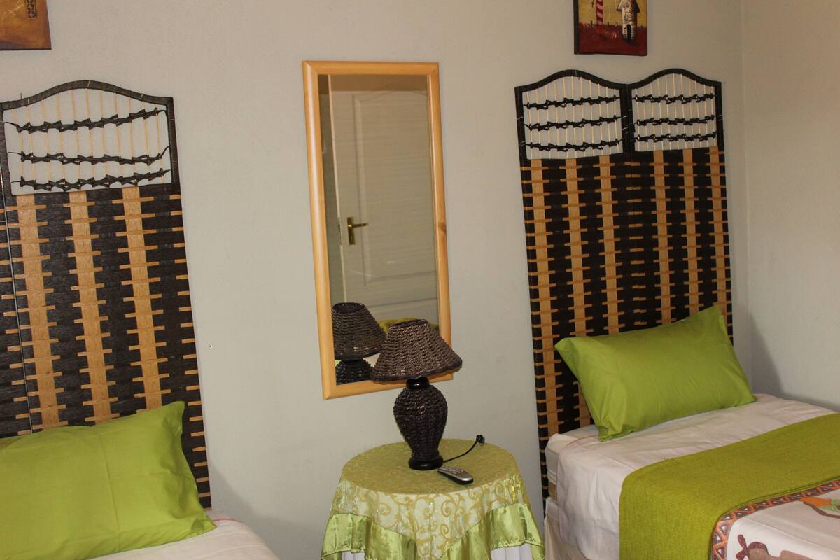 Una habitación con dos camas, cada una con una almohada verde y un cabecero estampado. Una mesa redonda con una lámpara y un espejo está en el centro.