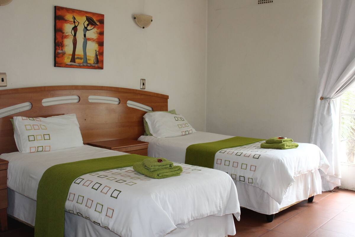 Una habitación de hotel con dos camas, cada una con ropa de cama blanca y detalles verdes, un cabecero de madera, un cuadro en la pared y una ventana con cortinas.