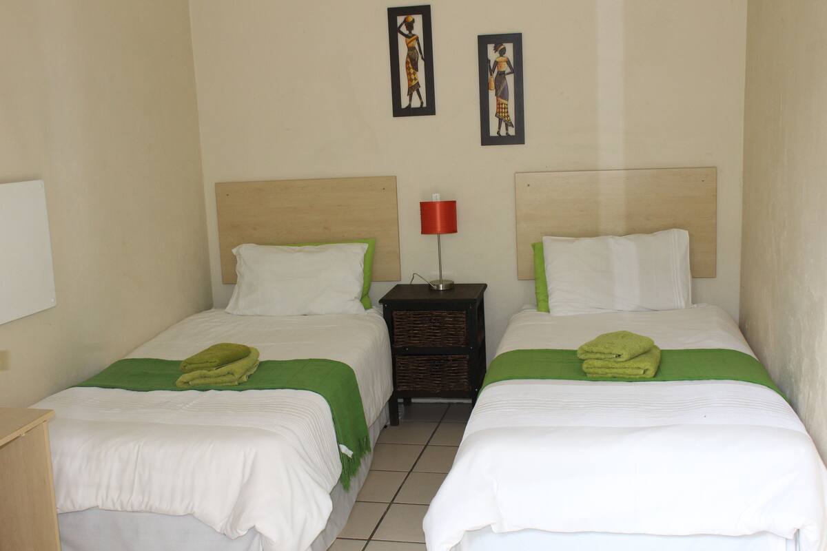 Habitación de hotel con dos camas, cada una con ropa blanca y detalles verdes, una mesita de noche con una lámpara roja, y una obra de arte en la pared.