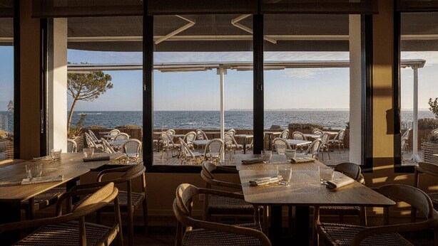 An indoor dining area with tables and chairs, large windows with a view of the sea and distant shoreline. - Logis Hôtel & Restaurant Les Mouettes - Larmor Plage, proche de Lorient (Larmor-Plage)