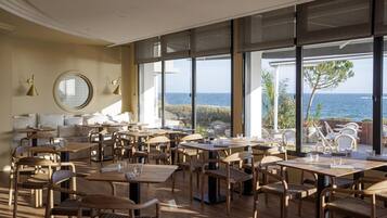 A spacious dining area with multiple tables and chairs, large windows offering a view of the sea, and a cozy seating area with cushions.