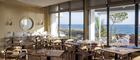 Un comedor amplio con varias mesas y sillas, ventanas grandes que ofrecen vista al mar, y un rincón de estar acogedor con cojines.