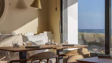 Un comedor moderno con un amplio ventanal con vista a la playa, muebles de madera y una lámpara colgante.