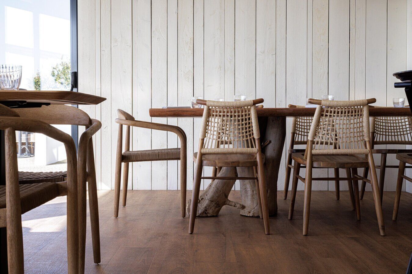 Un espace repas avec des tables et des chaises en bois, une grande fenêtre et un mur en bois peint en blanc.