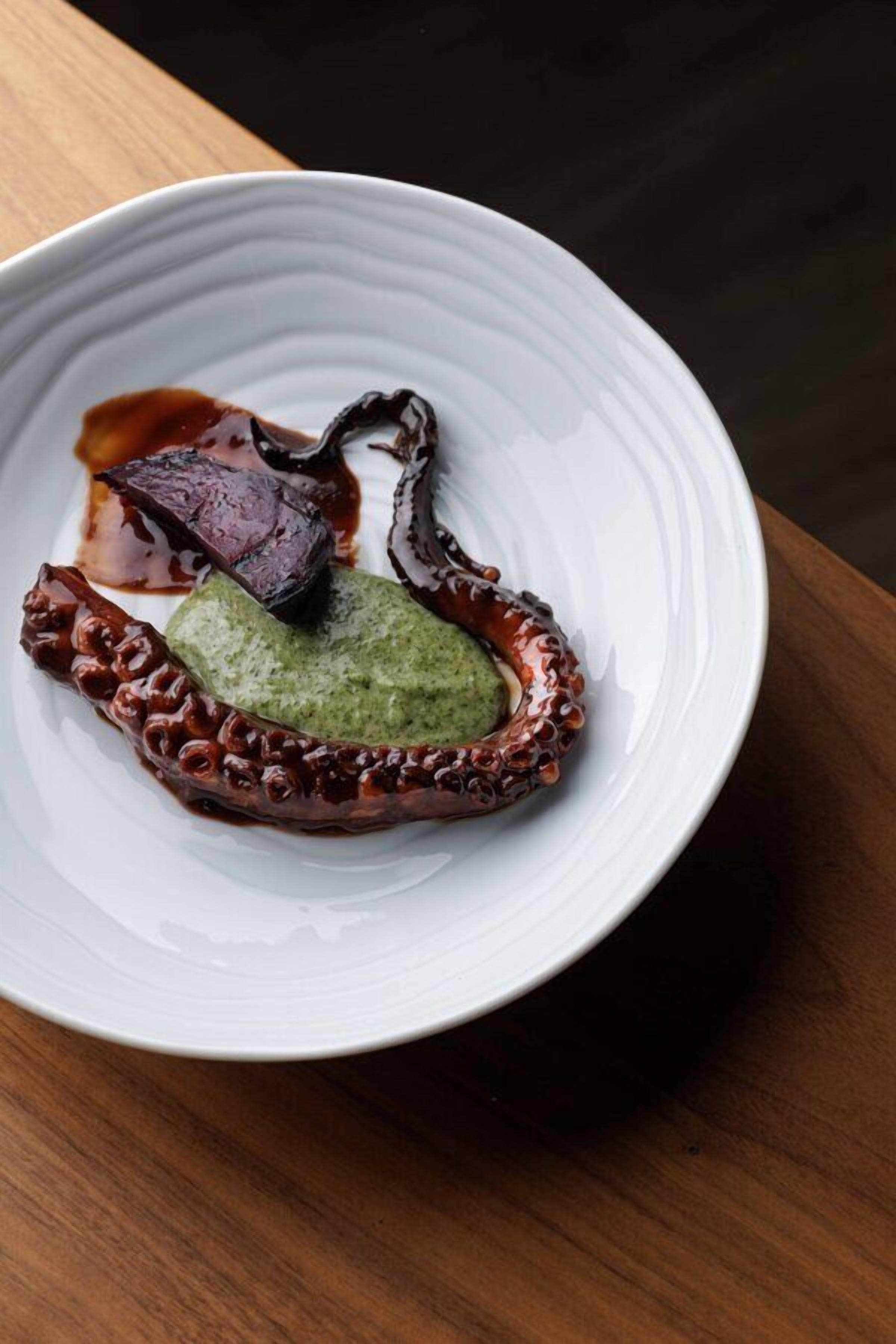 A white plate with octopus, green sauce, and brown sauce on a wooden table.