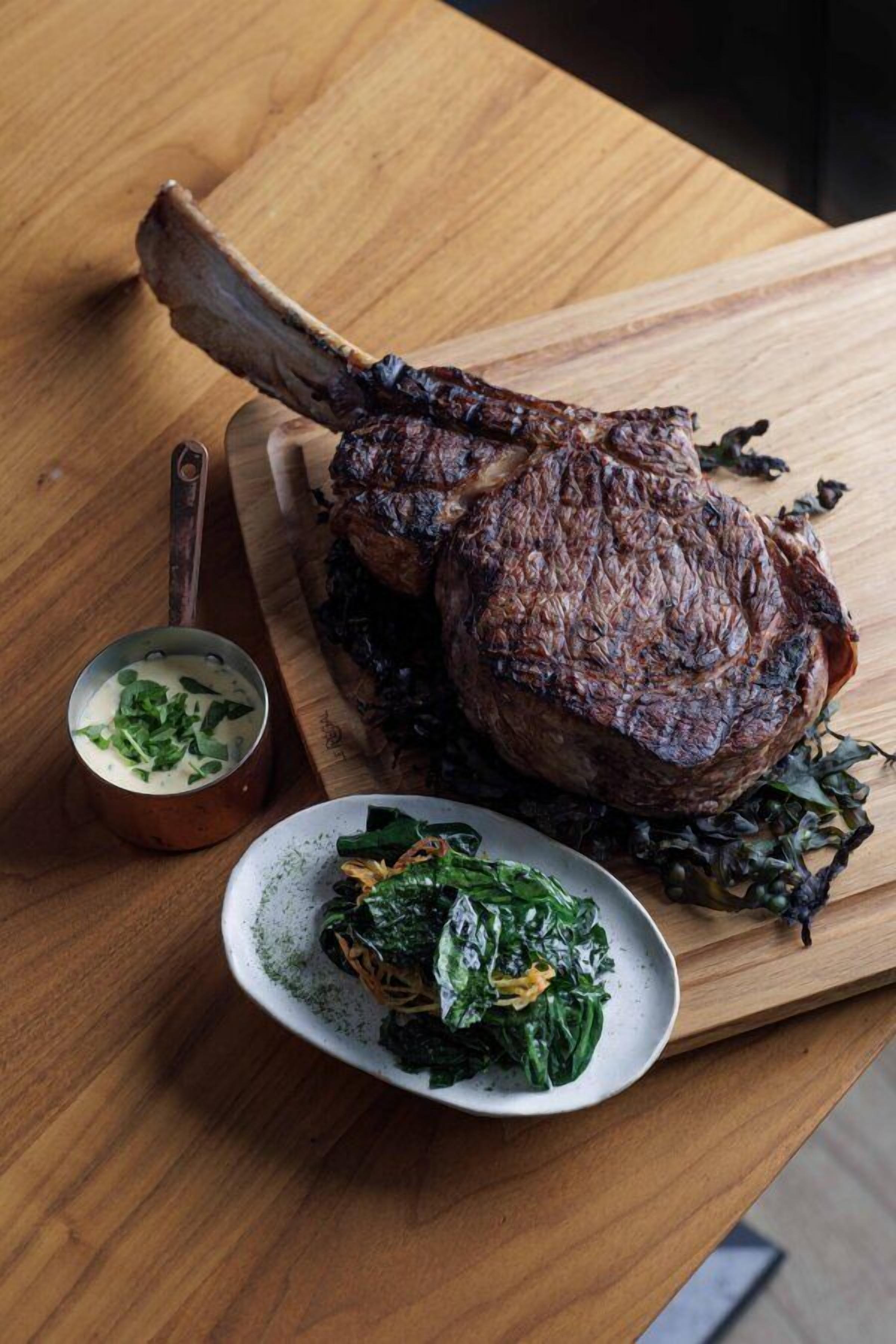 A roasted lamb leg on a wooden cutting board with a side of greens and a small bowl of sauce.