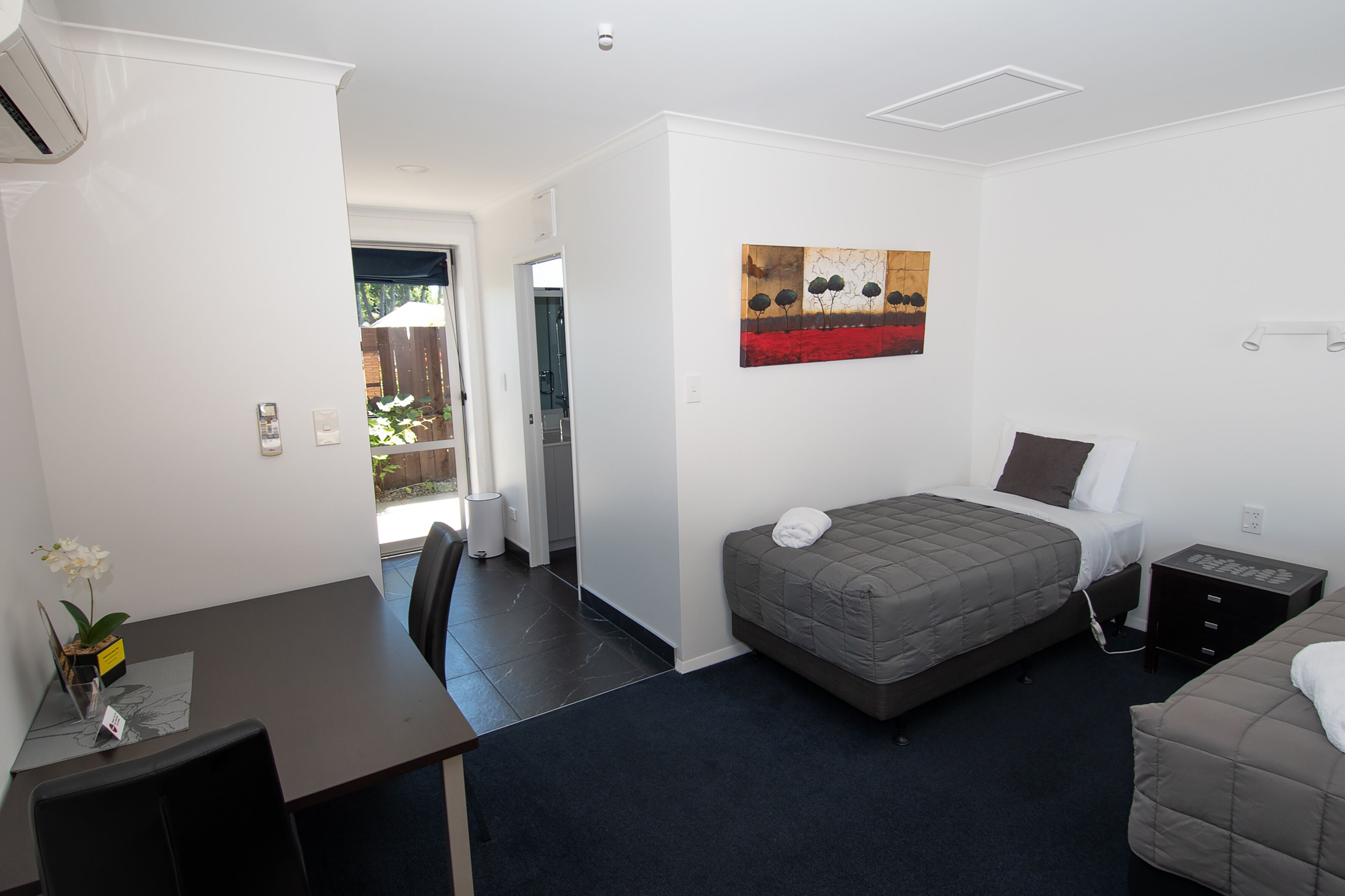 A compact room with a bed, bedside table, and a small desk with a chair. There is a window providing natural light and a door leading to an outdoor area.