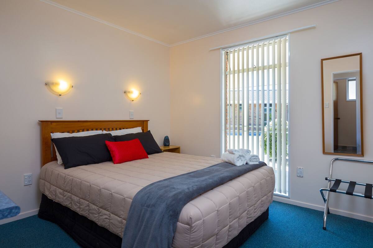 A bedroom with a bed, bedside tables, a window with blinds, a towel rack, and a mirror on the wall.
