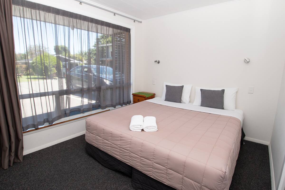 A hotel room with a large bed, a window with sheer curtains, a nightstand, and a view of a parking lot.
