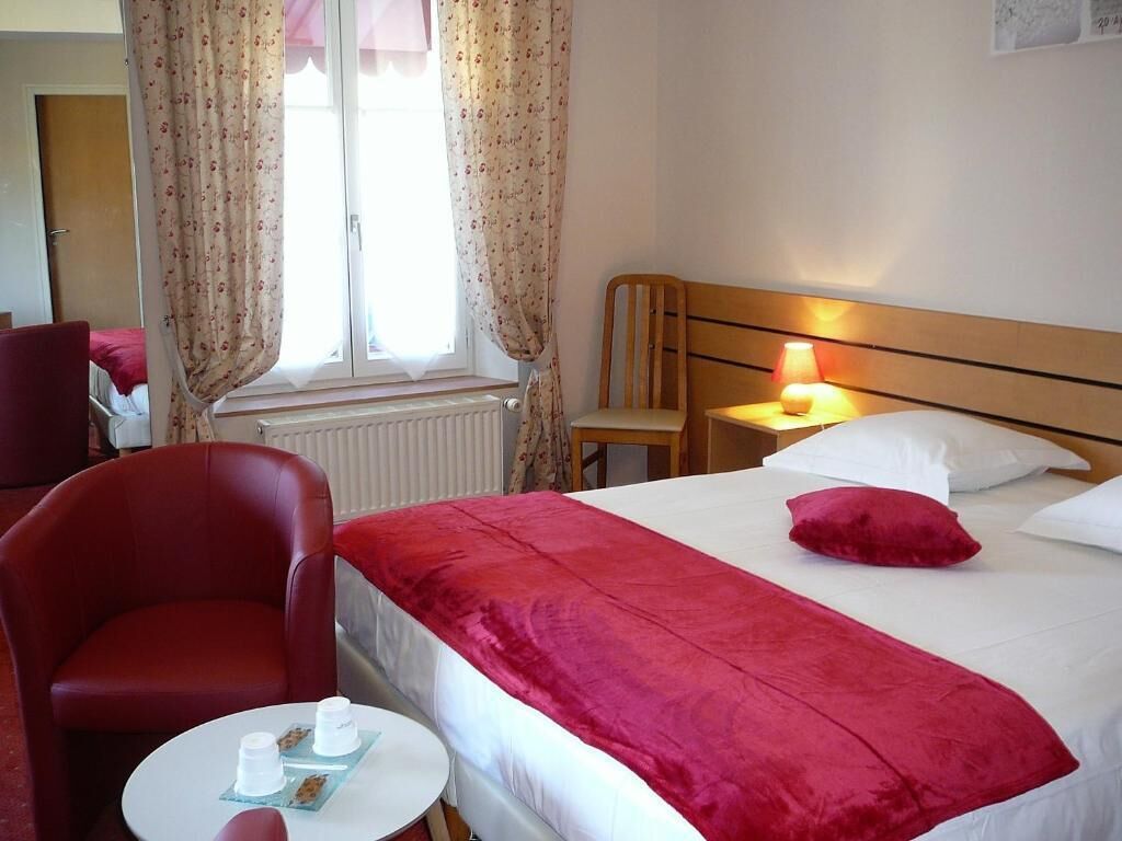 Une chambre d’hôtel avec un lit, une chaise, une table avec une lampe et une fenêtre avec des rideaux.