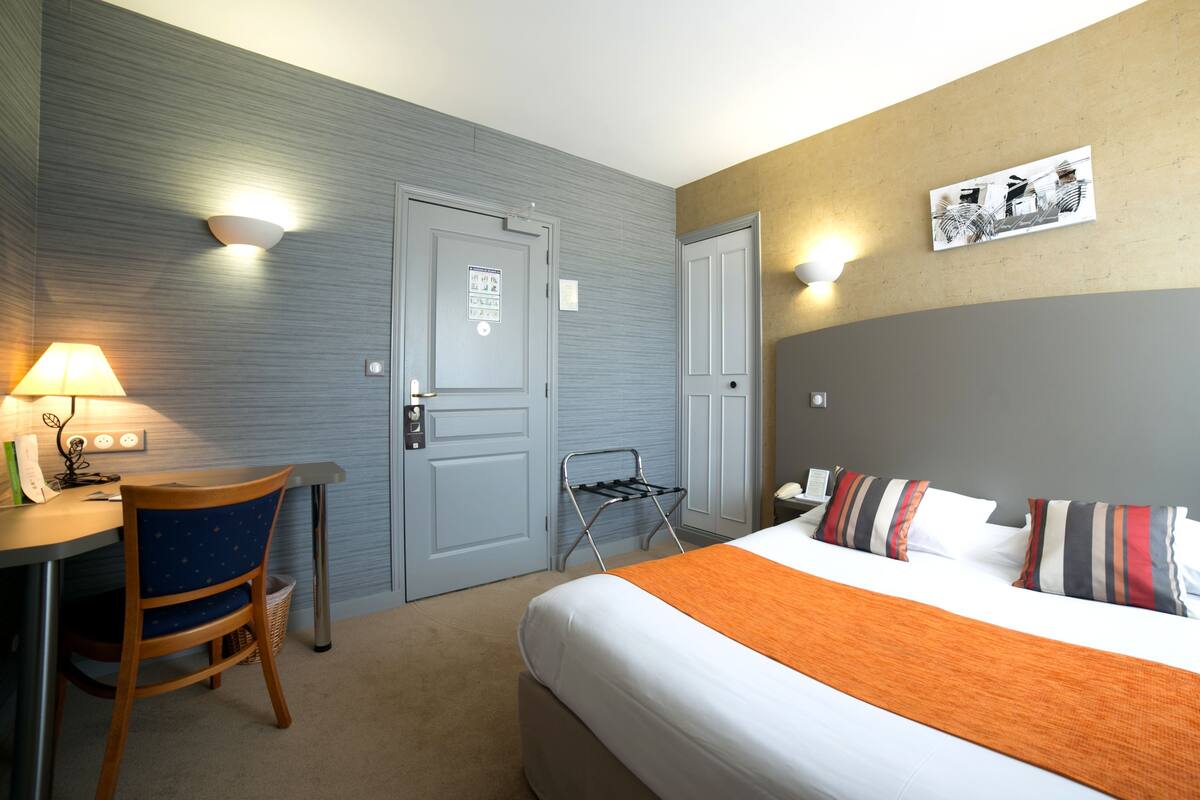 Ein Hotelzimmer mit Bett, Schreibtisch mit Lampe, Stuhl und einem an der Wand hängenden Kunstwerk.