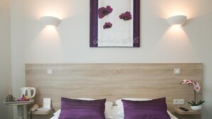 Ein Hotelzimmer mit einem hölzernen Kopfteil, zwei violetten Kissen, einem Nachttisch mit Telefon und Wasserkocher sowie einem Wandbild mit Blumenmotiven.