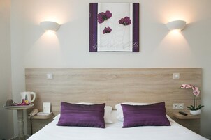 Habitación de hotel con cabecera de madera, dos almohadas moradas, una mesita de noche con un teléfono y una tetera, y una obra de arte floral colgada en la pared.