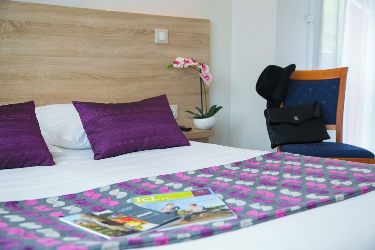 Hotelkamer met een houten hoofdeinde, een bed met wit en paars beddengoed, een stoel met een zwarte tas, een potplant en een tijdschrift op het bed.