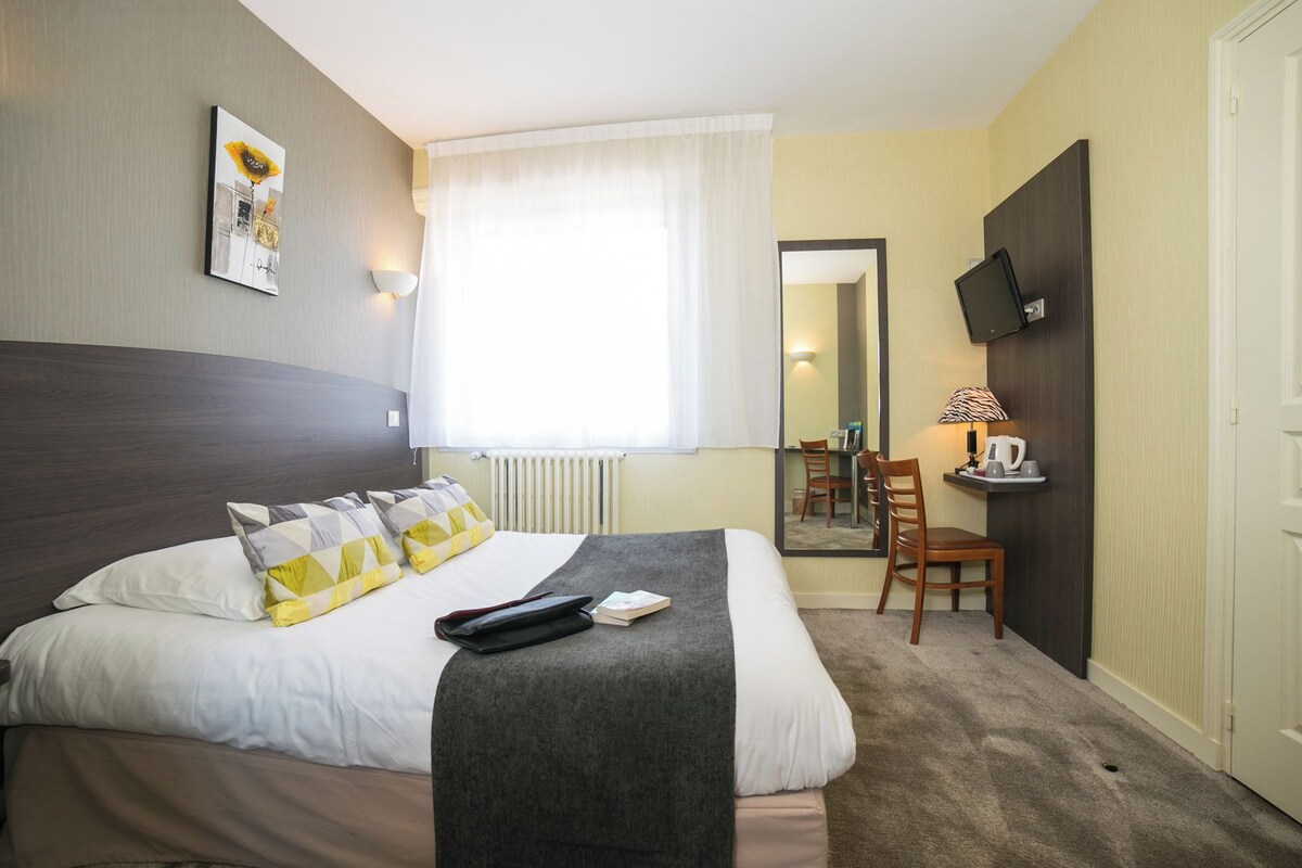 Hotellihuone, jossa on sänky, työpöytä tuolilla, seinälle asennettu televisio ja ikkuna, jossa on verhot.
