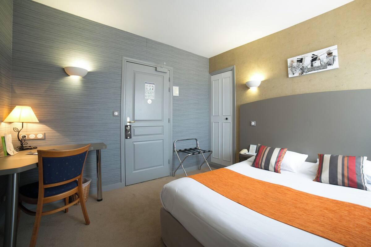 Ein Hotelzimmer mit einem Bett, einem Schreibtisch mit Lampe, einem Stuhl, einem Handtuchhalter an der Wand und einem Bild.