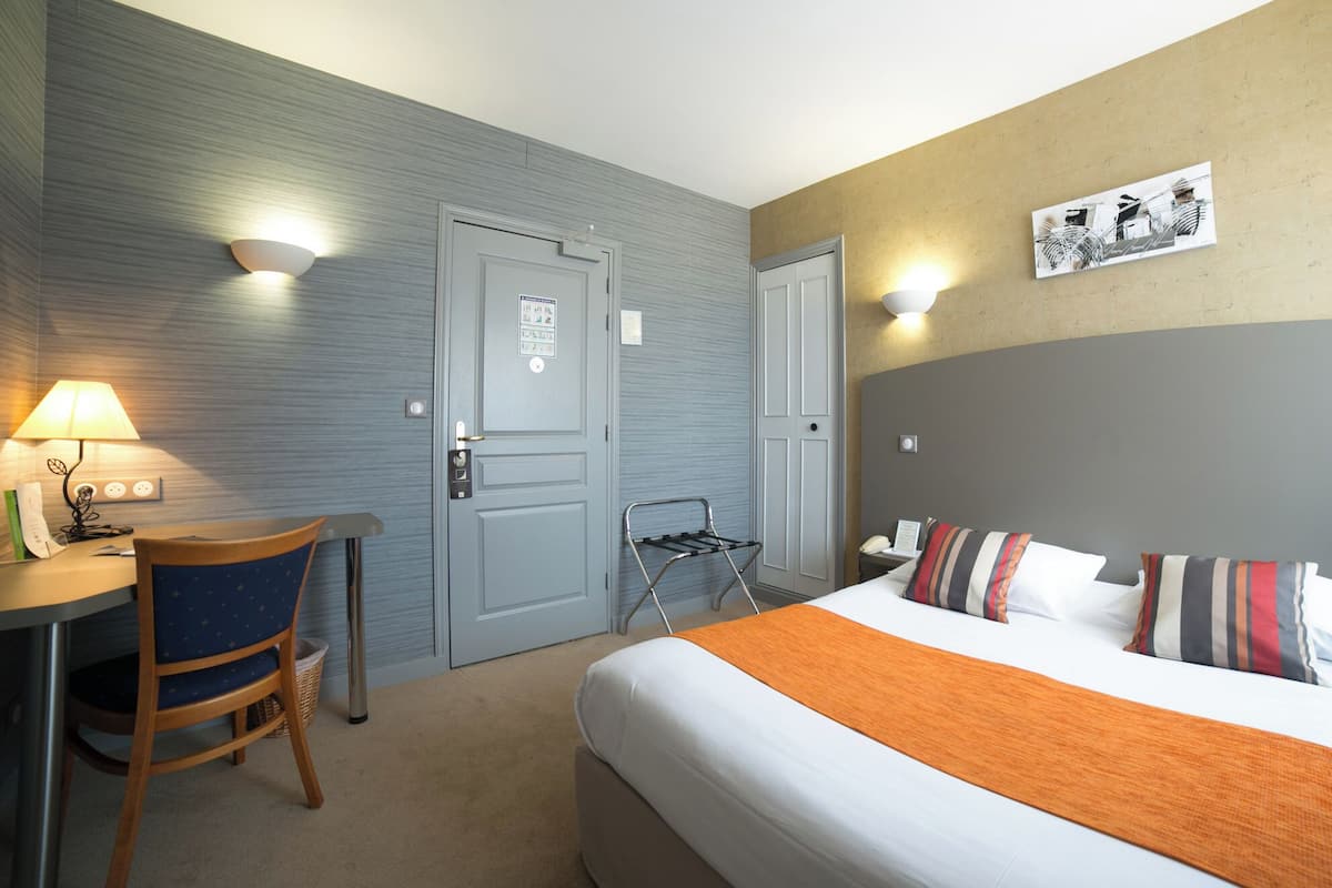 Une chambre d’hôtel avec un lit, un bureau avec une lampe, une chaise, un porte-serviettes fixé au mur et un tableau accroché au mur.