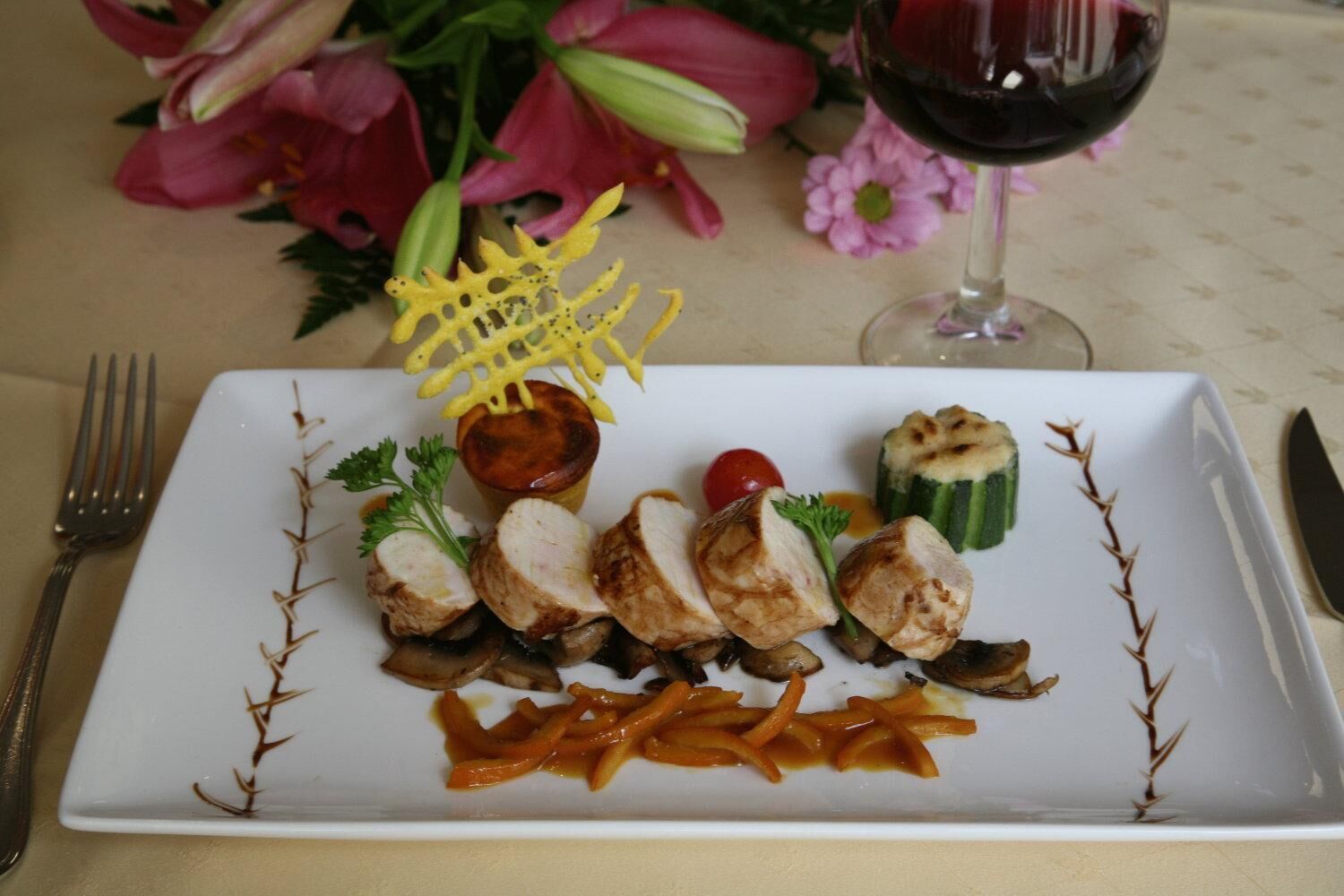 Ein weißes, rechteckiges Teller mit geschnittenem Fleisch, Pilzen, einer Kirschtomate und gegrilltem Zucchini, garniert mit Petersilie und einem gelben dekorativen Element.