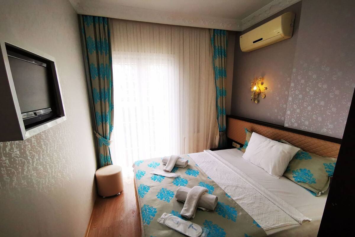 A hotel room with a bed, a TV mounted on the wall, a window with curtains, and an air conditioning unit.