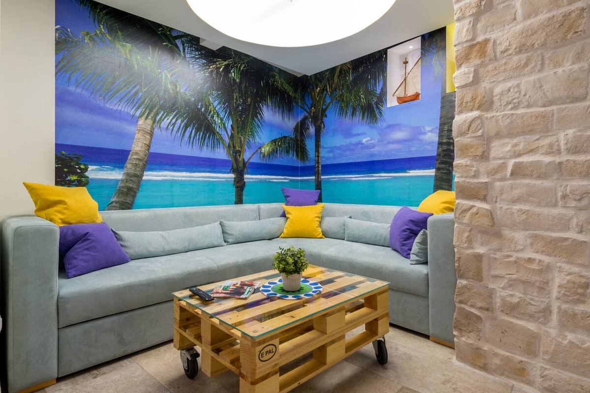 A room with a light blue sofa, yellow and purple cushions, a wooden coffee table with a plant, and a mural of a beach scene.