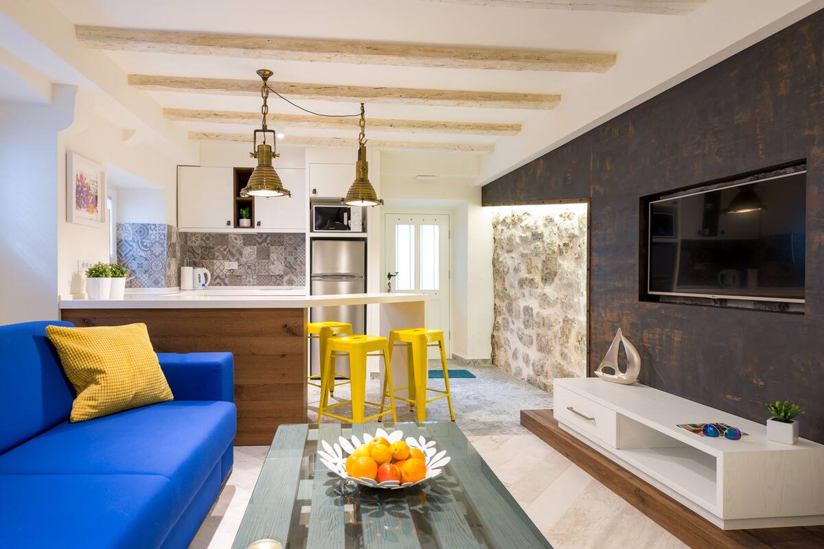 A modern living room with a blue sofa, yellow bar stools, a glass coffee table with fruit, and a TV on the wall.