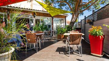 Un patio al aire libre con mobiliario de mimbre, plantas en macetas y un restaurante al fondo.