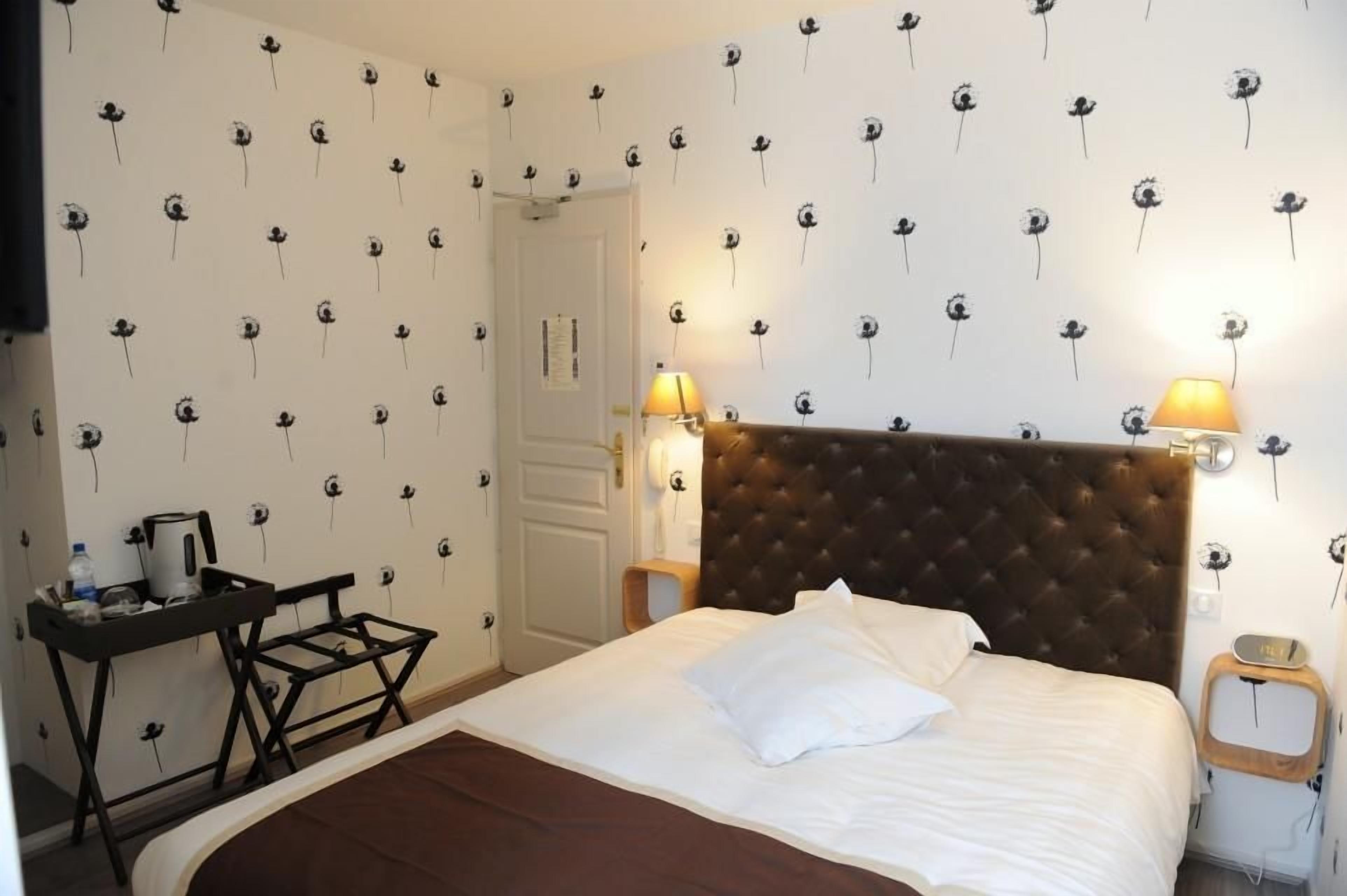 A hotel room with a bed, bedside lamps, a small table with a kettle, and a wall with a floral pattern.