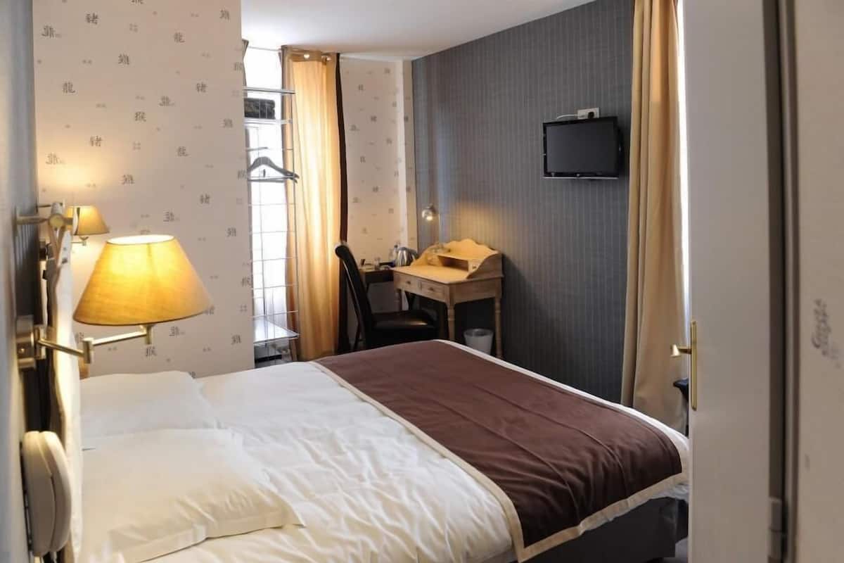 A hotel room with a bed, a desk, a chair, a TV mounted on the wall, and a window with curtains.