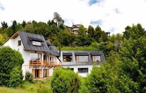Une maison à deux étages avec un balcon au premier étage, entourée d’une végétation luxuriante.