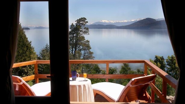 A wooden deck with a table and chairs overlooking a calm lake with mountains in the background. - La Sirenuse Lake Resort (San Carlos de Bariloche)