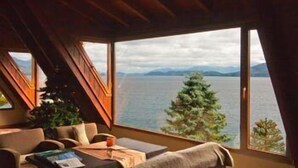 A room with a large window overlooking a lake and mountains, furnished with a sofa, a table, and a plant. - La Sirenuse Lake Resort (San Carlos de Bariloche)