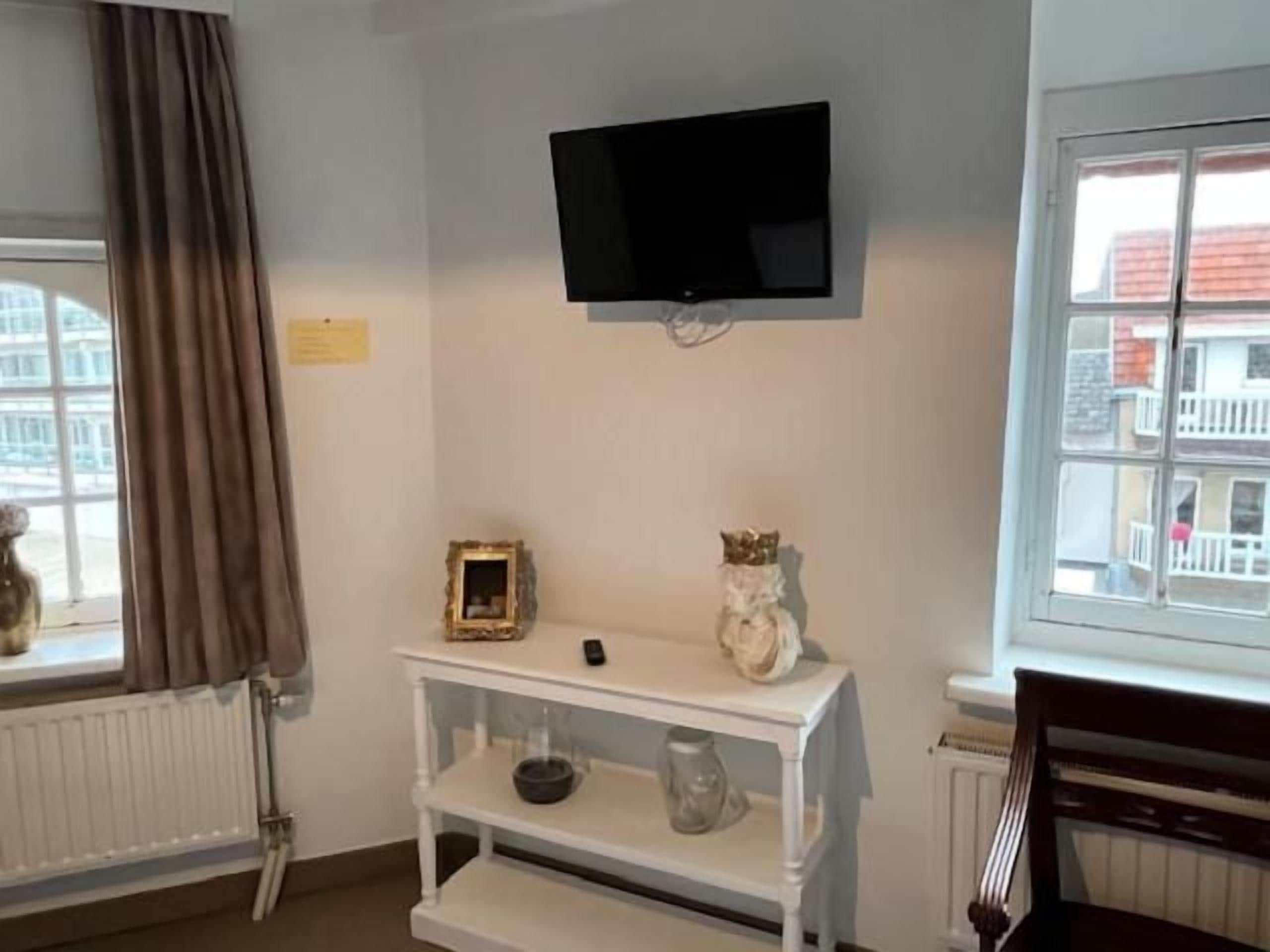 Una habitación con un televisor blanco montado en la pared, un estante blanco con adornos, una ventana con cortinas y un radiador.
