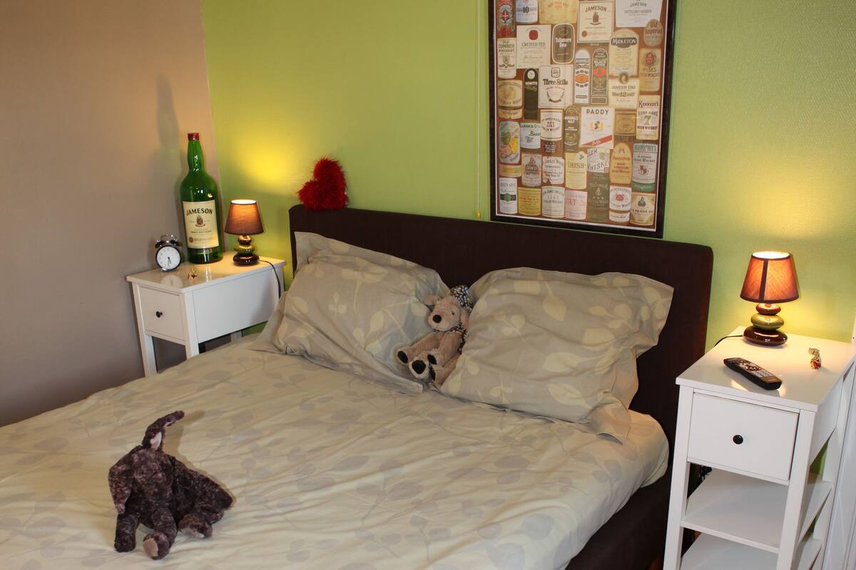 A neatly made bed with a teddy bear, two bedside tables with lamps, a wall clock, and a bulletin board.